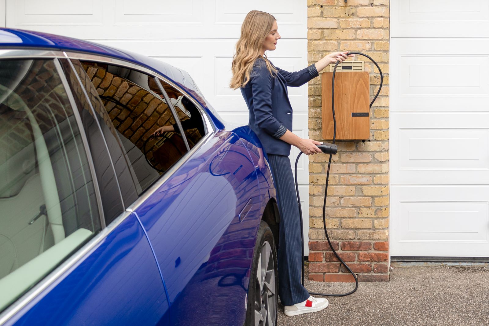 Kobieta w granatowym garniturze podłącza kabel do drewnianej ładowarki ściennej na ceglanym słupku obok niebieskiego samochodu elektrycznego