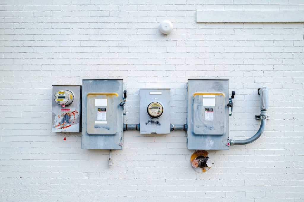Trzy szare skrzynki z licznikami energii elektrycznej i rurą osłonową na białej ceglanej ścianie